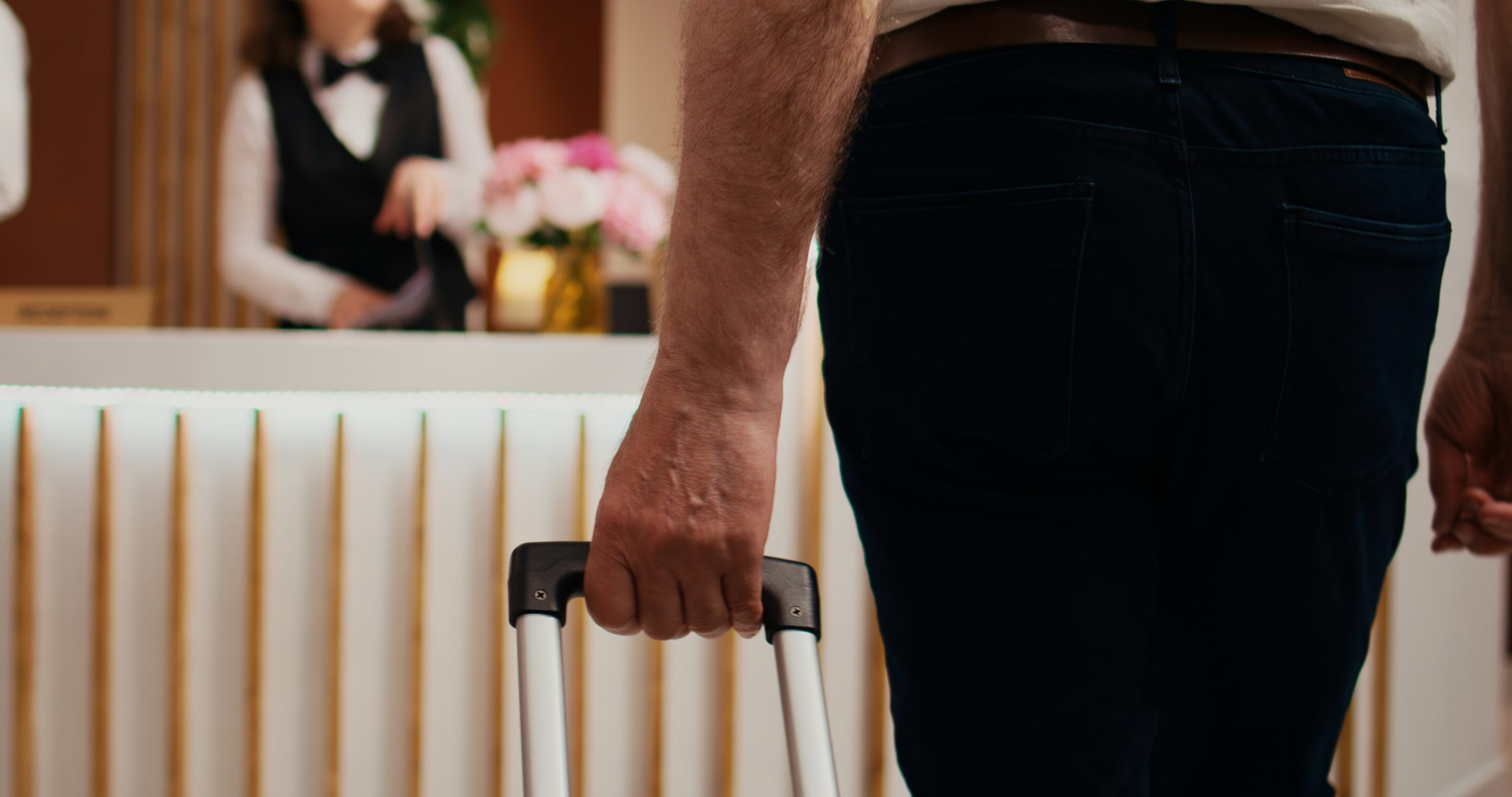 Zbliżenie od tyłu na mężczyznę w ciemnych spodniach trzymającego rączkę walizki. W tle, za ladą recepcyjną, widać pracownicę hotelu w czarnej kamizelce i muszce.