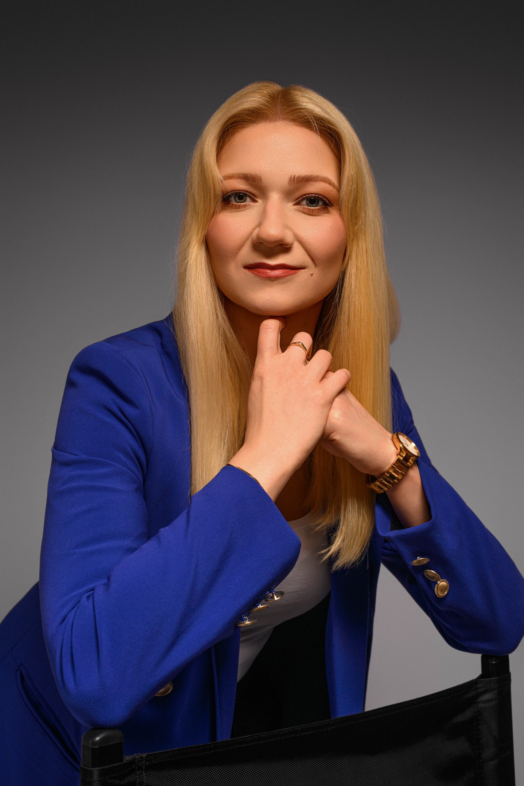 Portret uśmiechniętej kobiety z blond włosami, ubranej w niebieską marynarkę i białą bluzkę. Kobieta opiera się o oparcie krzesła, trzymając dłoń pod brodą. Tło jest jednolite, w szarym kolorze.